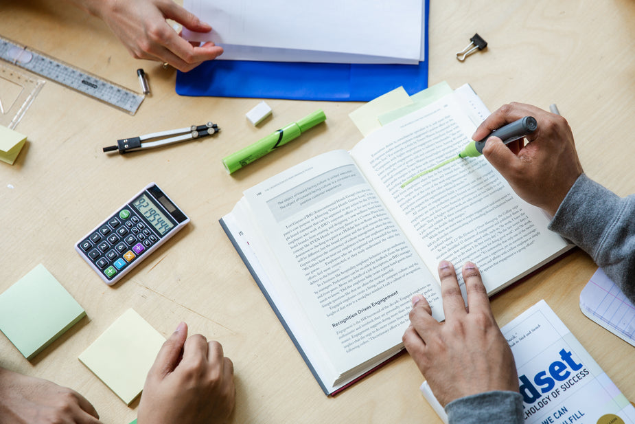 Study aids such as books, pens, note pads, and calculator on table with students' hands, photographed from above