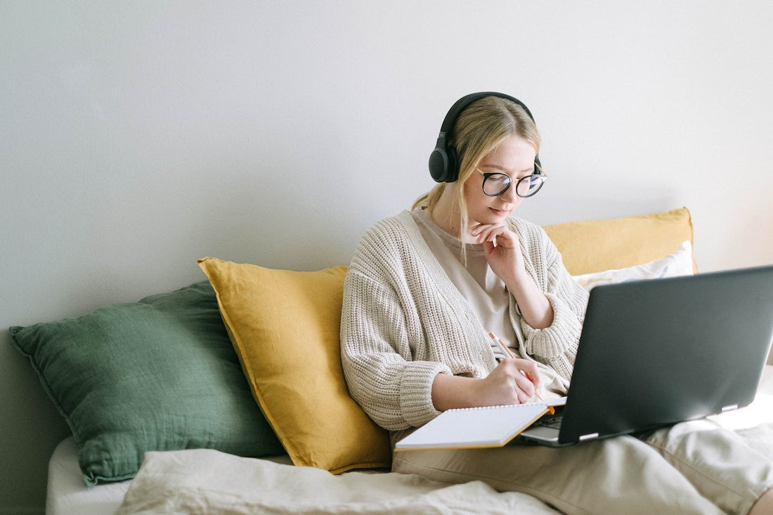 Woman on couch with laptop and headphones taking notes